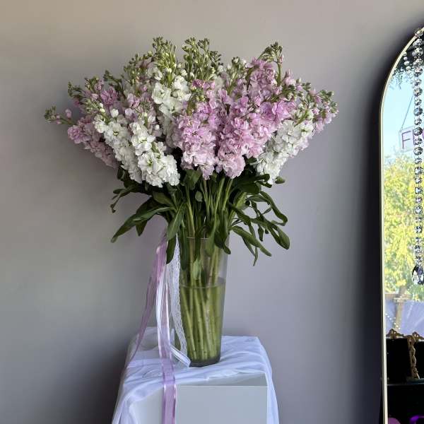 Tall arrangement of pink and white stock flowers in a clear glass vase with trailing ribbons