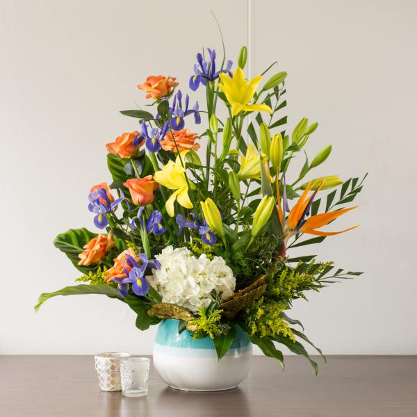 Tall mixed bouquet with yellow lilies, blue irises, orange roses, and white hydrangea in a round vase