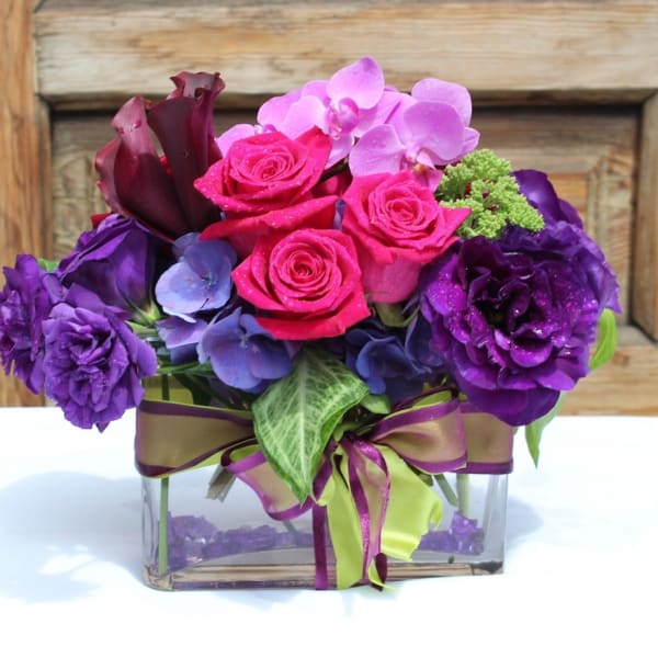 Purple and pink floral arrangement in a clear square vase with ribbon