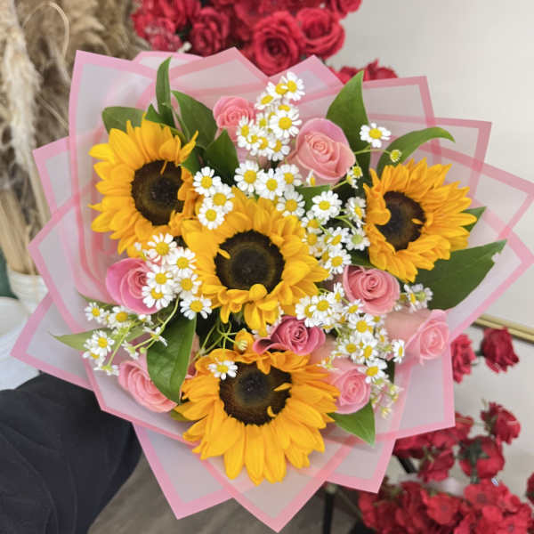 Hand-tied bouquet of yellow sunflowers, small white daisies, and pink roses wrapped in layered pink paper.