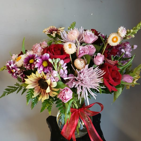 Mixed bouquet of roses, chrysanthemums, and sunflowers in a glass vase with a red ribbon