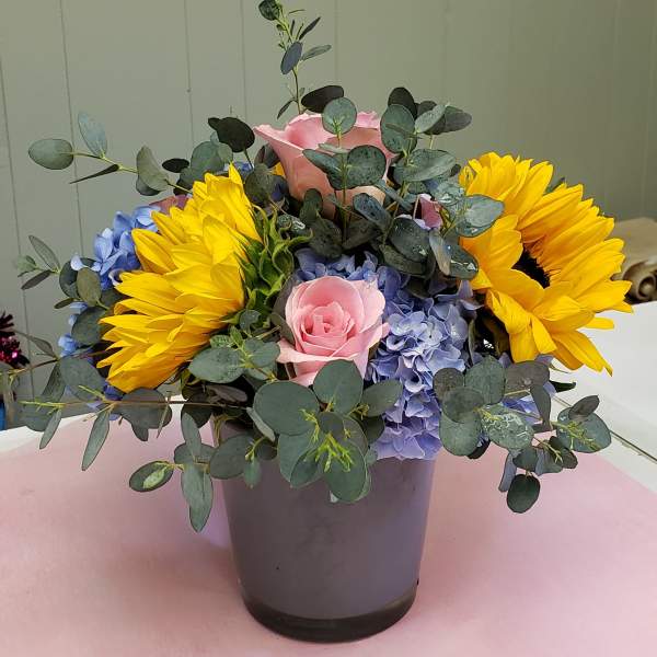 Arrangement of yellow sunflowers, pink roses, and blue hydrangeas in a gray vase