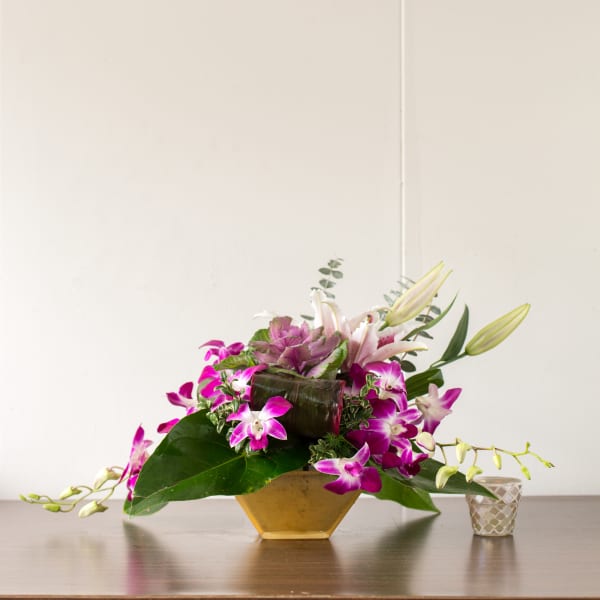 Purple orchids and lilies arranged in a gold vase with a small candle holder beside it