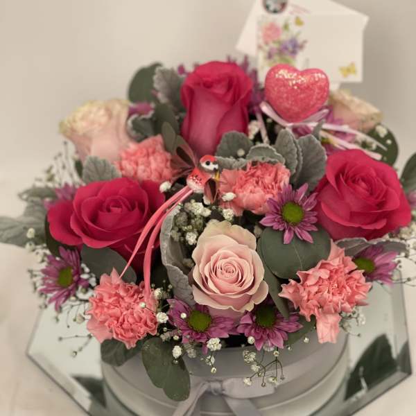 Pink and red roses with carnations in a round box, accented by a heart pick