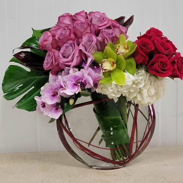 Low glass vase arrangement with lavender roses, red roses, green orchids, white hydrangeas, and purple orchids.