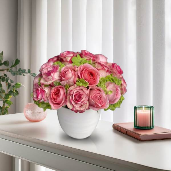 Pink roses arranged in a white vase on a table beside a candle and book