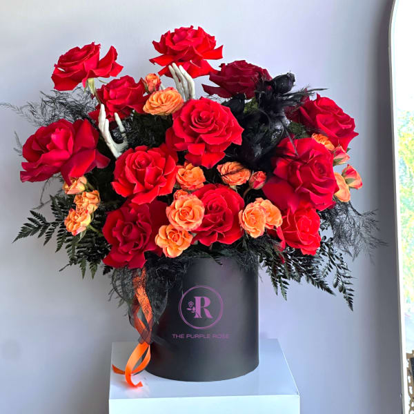 Hatbox arrangement of red roses and orange spray roses with black accents and skeleton hand decorations.