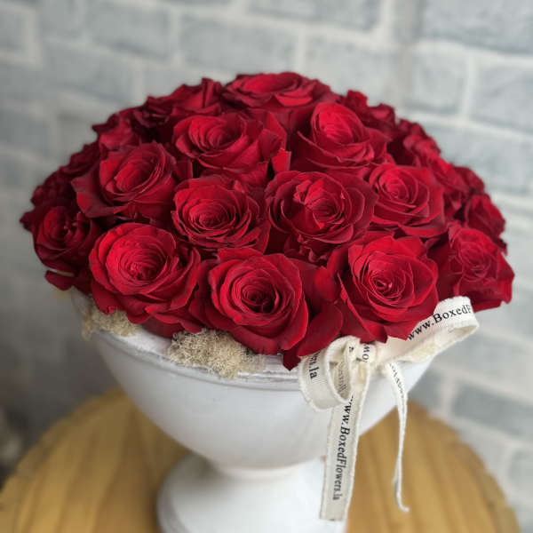 Red roses arranged in a white pedestal vase with a ribbon bow