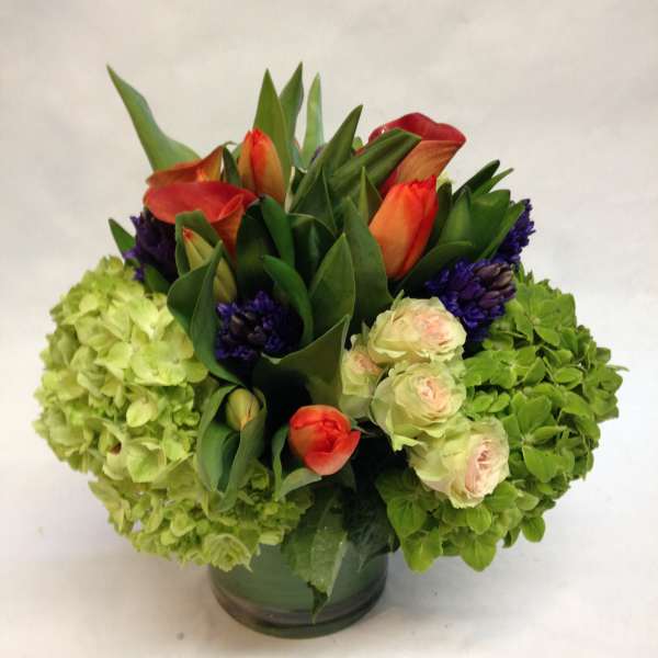 Compact arrangement of green hydrangeas, orange tulips, cream roses and purple blooms in a round glass vase