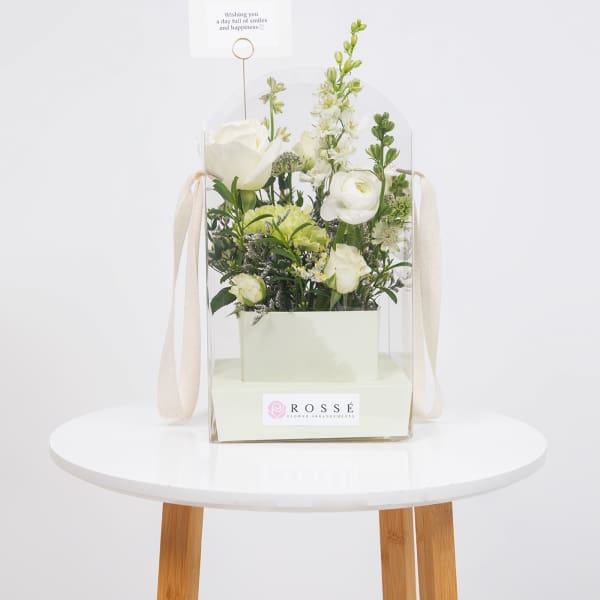 White floral arrangement in a clear gift box with a small card topper