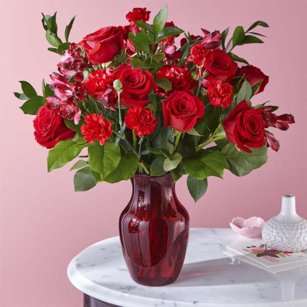 Arrangement of red roses and carnations in a red glass vase on a marble table.