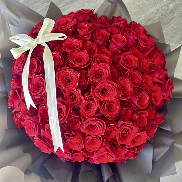 Round bouquet of red roses wrapped in dark paper with a white ribbon