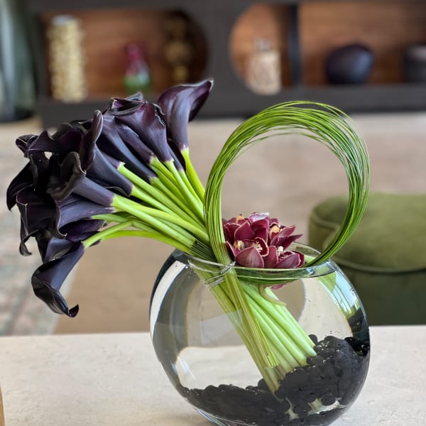 Purple calla lilies and a small burgundy orchid arrangement in a round glass vase