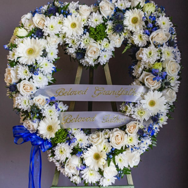 Heart-shaped floral wreath with white flowers and blue accents on a stand