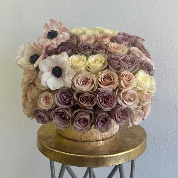 Round bouquet of blush, lavender, and cream roses with pale anemones in a gold box