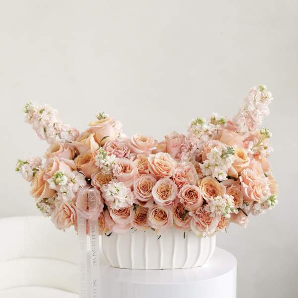 Peach rose arrangement in a white vase with trailing ribbon