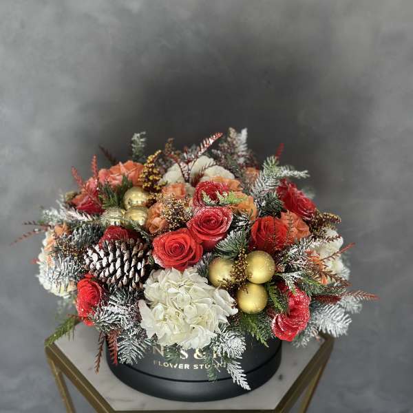 Round floral arrangement with red roses, white hydrangea, pinecones, and gold ornaments in a black box