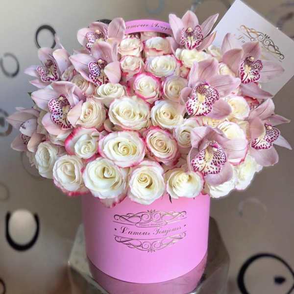 Round pink hat box filled with cream roses edged in pink and pale pink orchids, with a small card tucked in.