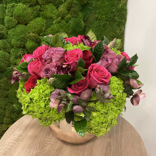 Low round arrangement of hot pink and mauve roses with green hydrangeas in a metallic bowl vase