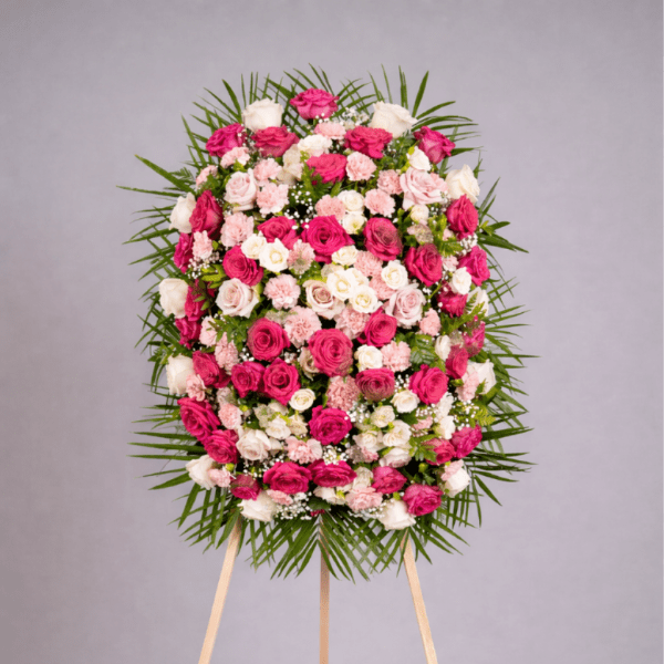 Standing oval spray of pink and white roses and carnations on an easel, framed by a ring of fanned greenery.