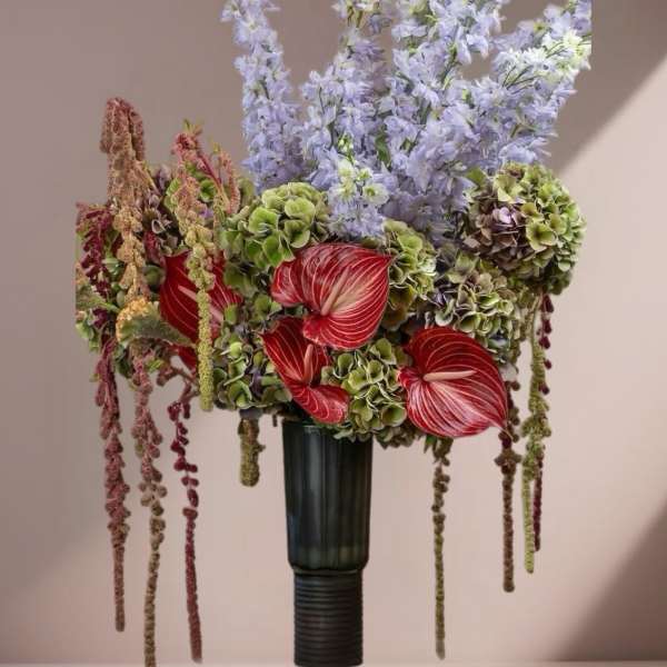 Tall arrangement with red anthuriums, green hydrangeas, and pale lavender flowers in a dark vase.