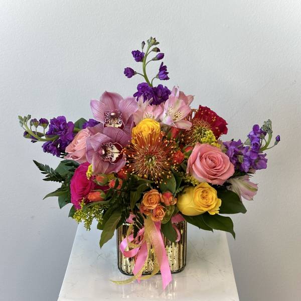 Colorful bouquet of roses, orchids, and mixed blooms in a gold vase with pink ribbon
