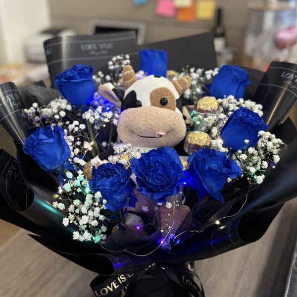 Blue roses arranged around a plush cow with baby’s breath and chocolates