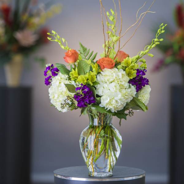 Mixed bouquet of white hydrangeas, orange roses, and purple flowers in a glass vase