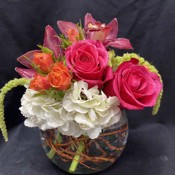 Short arrangement of hot pink roses, orange spray roses, orchids and white hydrangea in a glass bowl vase