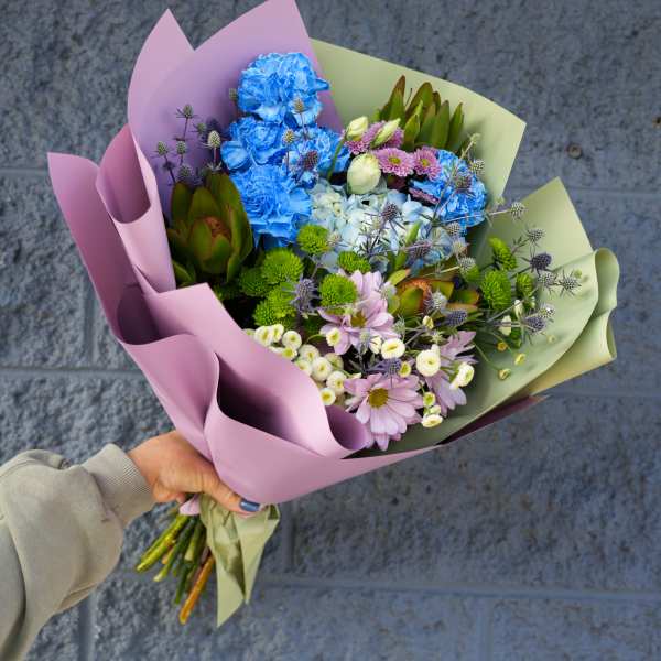 Handheld bouquet of blue, pink, and white flowers wrapped in pastel paper
