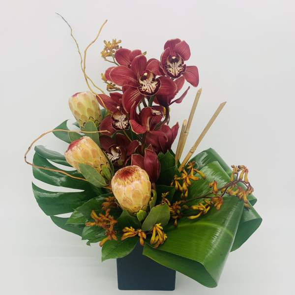 Tropical arrangement with red orchids and pale protea in a black cube vase with bold green leaves.