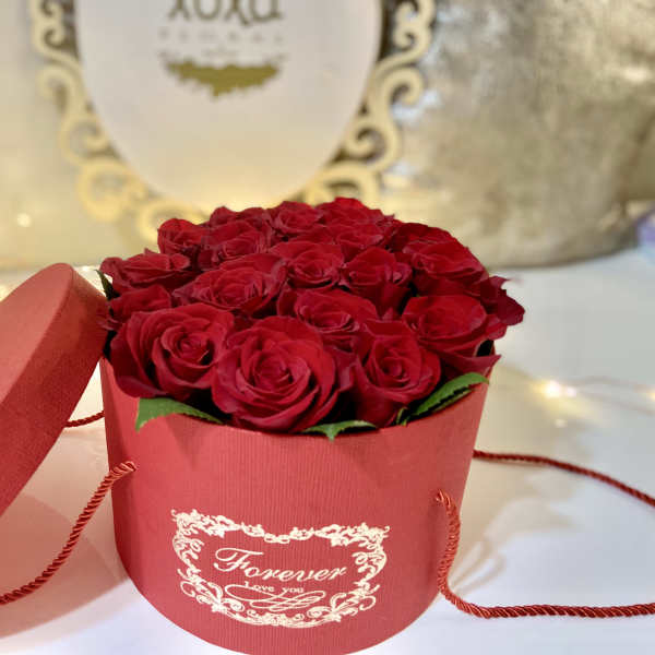 Red roses arranged in a round red gift box with a lid beside it.