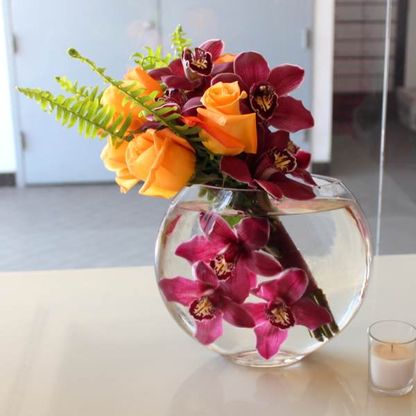 Orange roses and burgundy orchids in a round glass vase