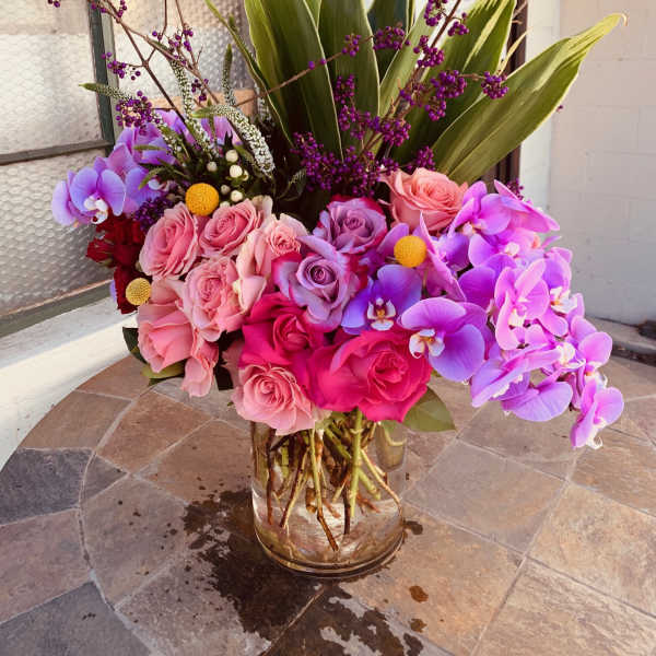 Pink roses and purple orchids arranged in a glass vase with tall green leaves.