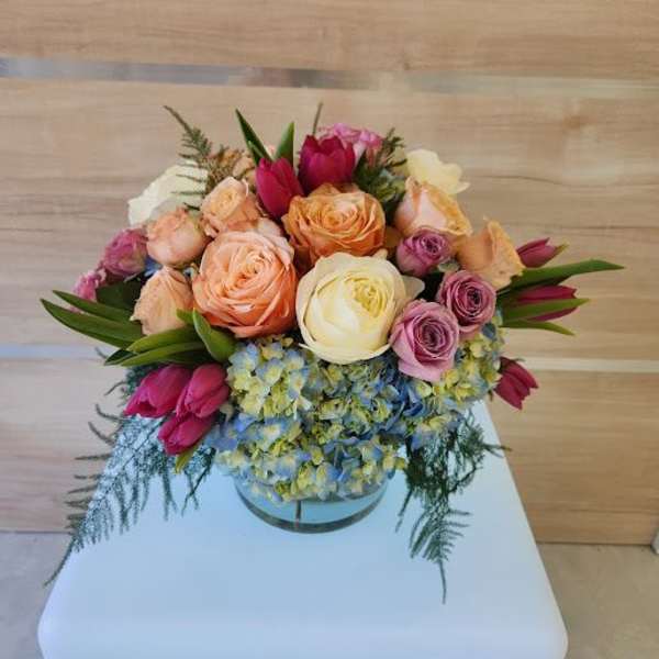Short round arrangement of peach and cream roses, pink tulips, and blue hydrangeas in a clear glass vase
