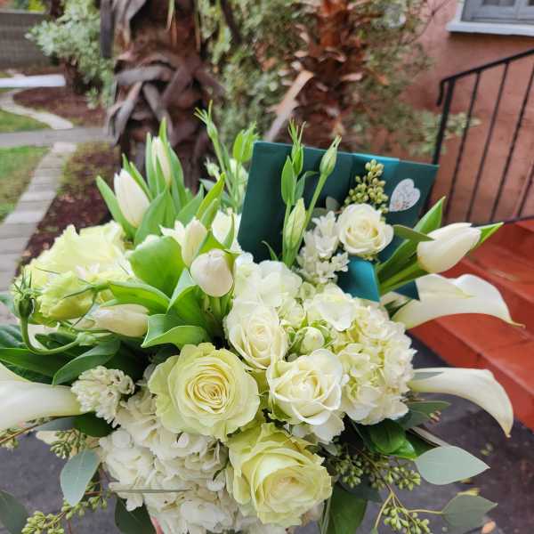Low arrangement of white roses, hydrangeas, tulips, and calla lilies in a clear glass vase with a green card.