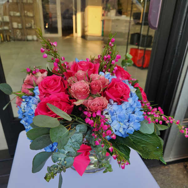 Low glass vase arrangement with hot pink roses and blue hydrangeas accented by small pink blossoms.