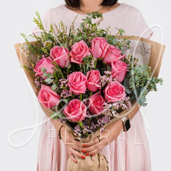 Bouquet of bright pink roses with tiny pink filler flowers wrapped in kraft paper, held by a person in a pale dress.