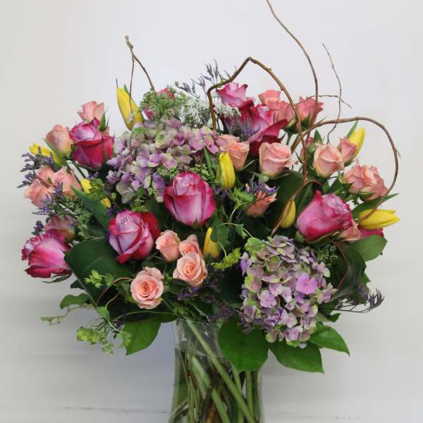 Tall mixed arrangement of pink roses, peach spray roses, yellow tulips, and purple hydrangeas in a glass vase