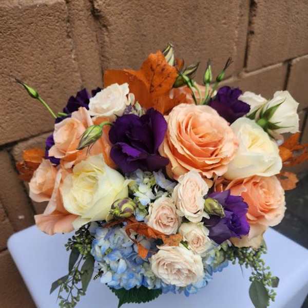 Compact arrangement of peach and cream roses, purple lisianthus, and blue hydrangea with orange leaves
