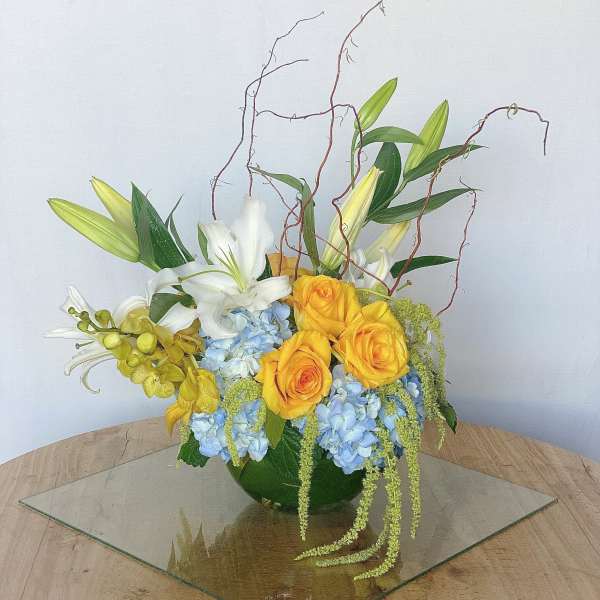 Arrangement of yellow roses, white lilies, and blue hydrangeas in a green vase