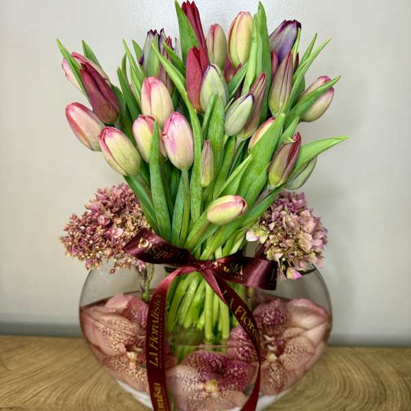 Pink and purple tulips in a round glass vase with a burgundy ribbon