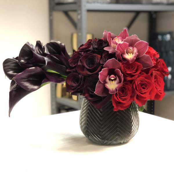 Low arrangement of dark purple calla lilies, red roses, and mauve orchids in a textured dark vase
