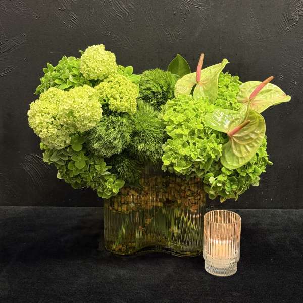 Green floral arrangement in a glass vase with an anthurium and candle
