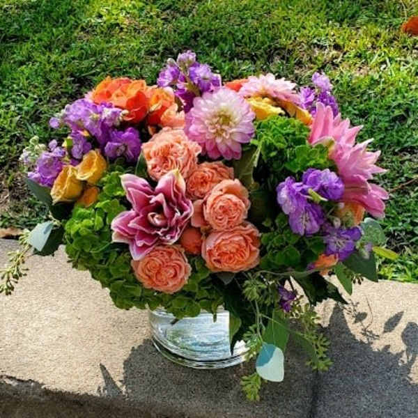 Low mixed bouquet of peach roses, pink lilies, and purple flowers in a clear glass vase outdoors