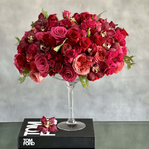Tall glass compote filled with dense red and hot pink roses and orchids on a black book.