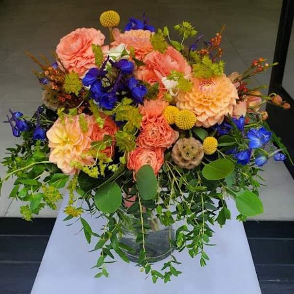 Large peach and blue mixed flower arrangement with trailing foliage in a clear glass vase.