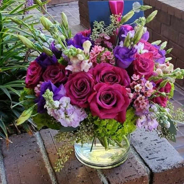 Low glass vase of fuchsia roses, purple blooms, and mixed pink flowers with a card tucked at the back.