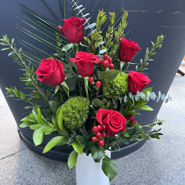 Red roses arranged in a white vase with greenery and red berries