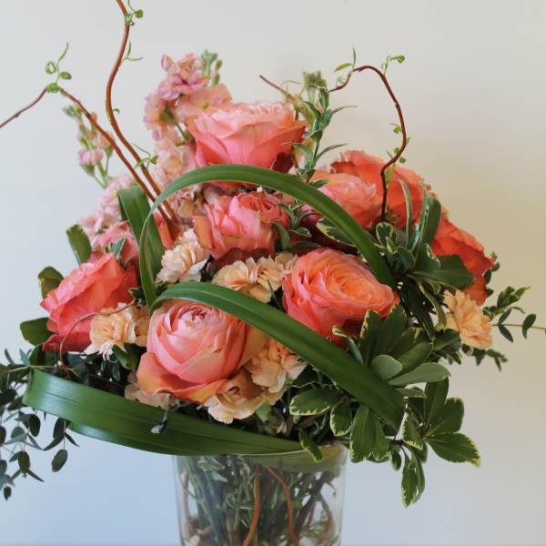 Tall arrangement of coral roses and peach carnations with looping greens in a clear glass vase